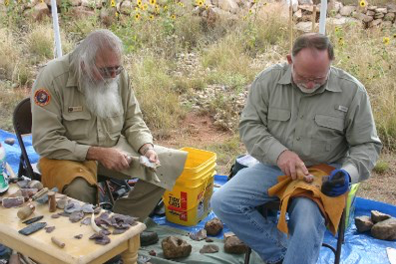 In a field, two gentlemen demonstrate knapping next to a table of stones
