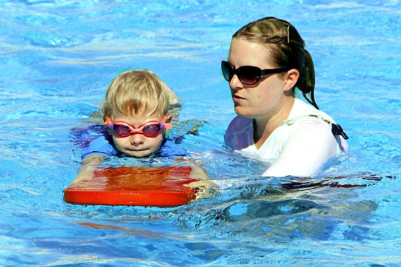In the water and using a red kickboard, a ponytailed blonde in a white T is teaching a blond toddler in a blue T how to swim