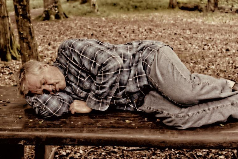 A blond-haired man in a plaid flannel shirt lying on his side on a park bench