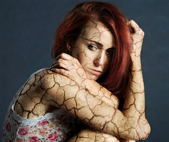 Close-up of a woman with dark red hair, curled up with her elbows on her knees, and cracks showing all over her body