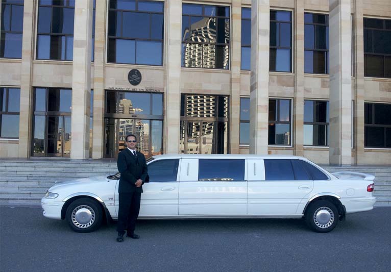 A chauffeur standing in front of a stretch limo in front of a hotel.