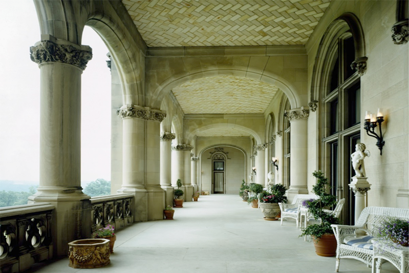 A very wide porch of limestone blocks and herringbone-tiled ceiling and arches overlooking a view. It's furnished with groupings of wicker, stone planters, and statues.