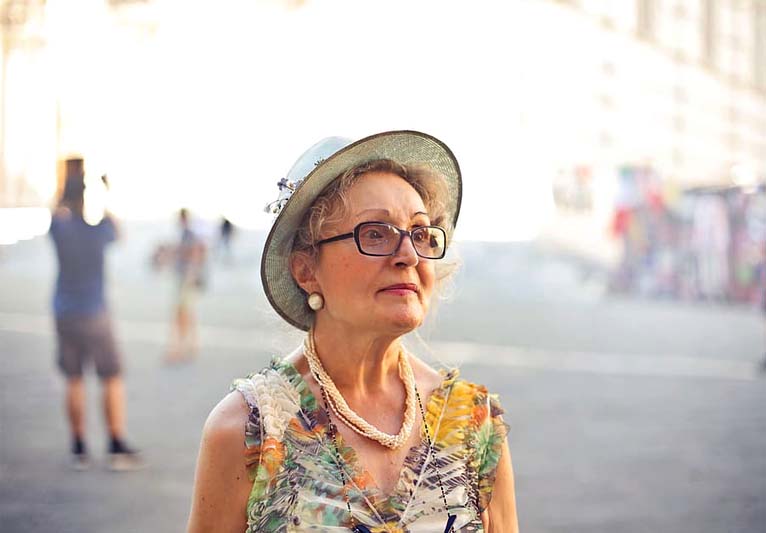 A soft pastel background showcases an older woman in a sunhat and glasses.