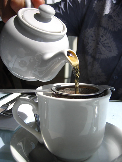 A close-up of a white teapot pouring tea into a strainer situated on topo f a white cup.