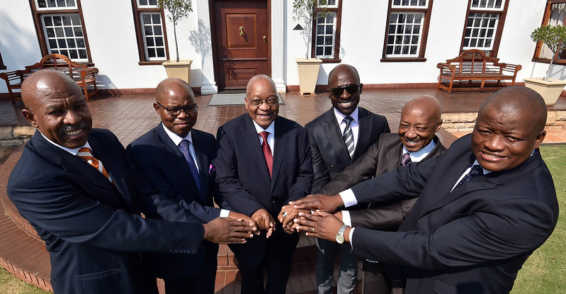 Outside a house, politicians stand in a semi-circle clasping hands