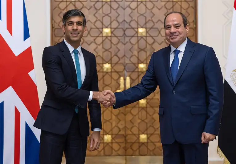 The British Prime Minister and the Egyptian President face forward while shaking hands, their country's flags on either side of them.