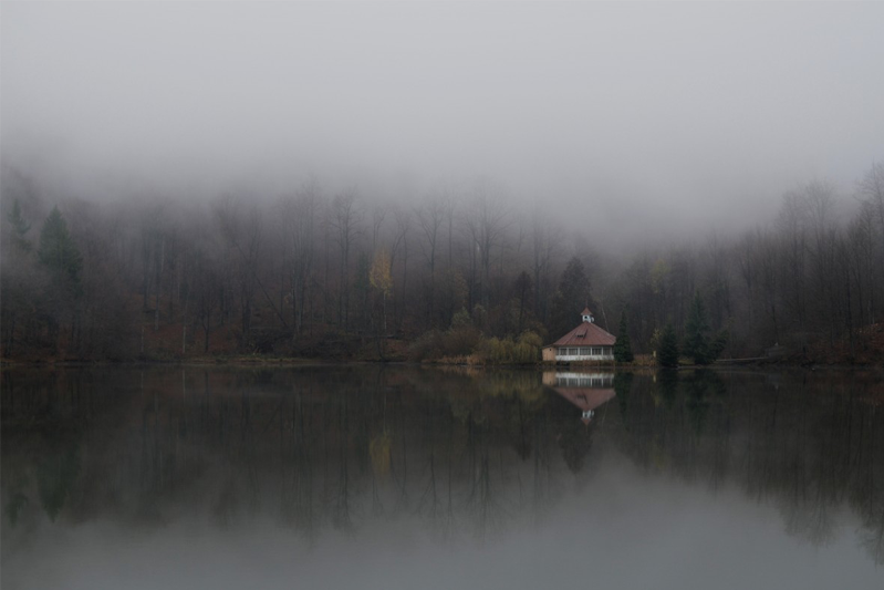 A house in the distance reflected in the lake with the mist covering the forest.