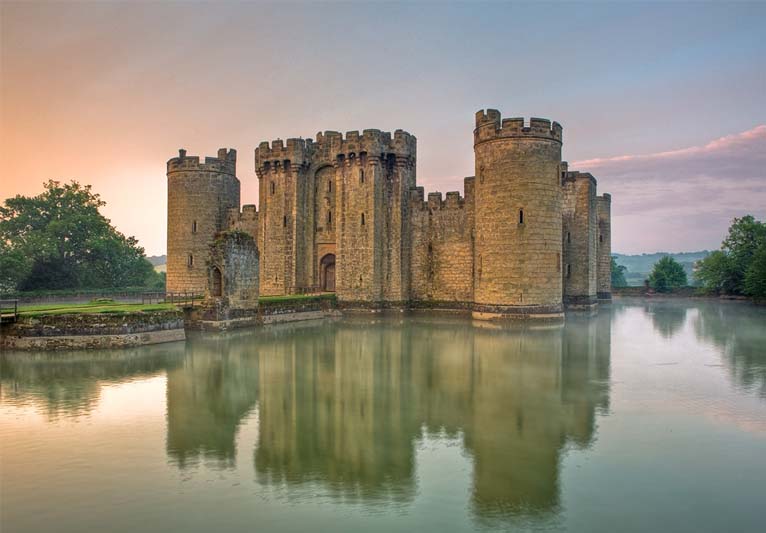 Sunrise creates a soft glow around a castle fronted by a wide moat.