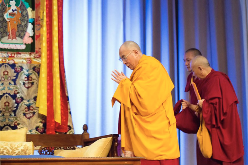 The dalai lama in a yellow robe with two acolytes in red bow to the left