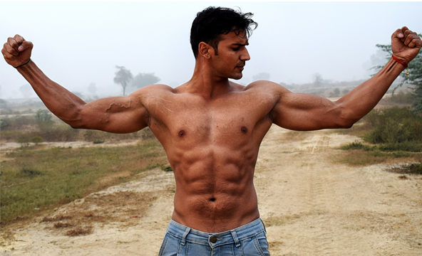 A young shirtless man in jeans standing on a dirt road, flexing his muscles