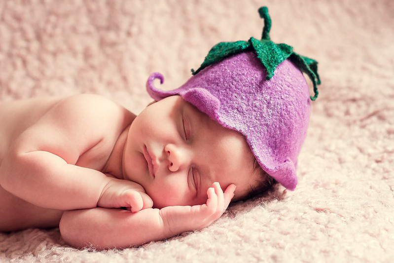 The top half of a cute baby lying on a sherpa blanket and wearing a lavender knit cap that looks like an upside down flower with a green stem emerging from the top of the cap.