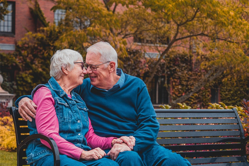 Older couple dressed casually are embracing on a park bench against a background of autumnal trees