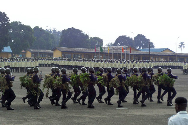 A unit of Navy SEALS march past large groups of sailors in cream-topped uniforms.