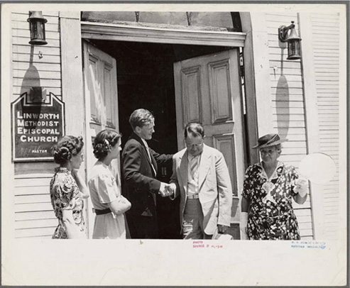 A black-and-white photo of a pastor shaking hands with a besuited man in the doorway of a white clapboard church. Two women are standing in profile to the left.