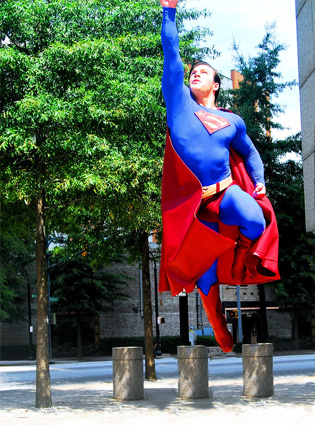 Superman lunging up from the ground, his right arm thrust upward and his left leg bent with trees and buildings in the background.