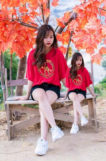 Woman with long brown hair wearing a red T-shirt and black shorts is sitting on a wooden bench with a red maple tree behind her. Her petite twin sits next to her.