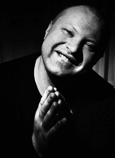 Black background with a close-up of a man smiling with his hands in a prayerful pose