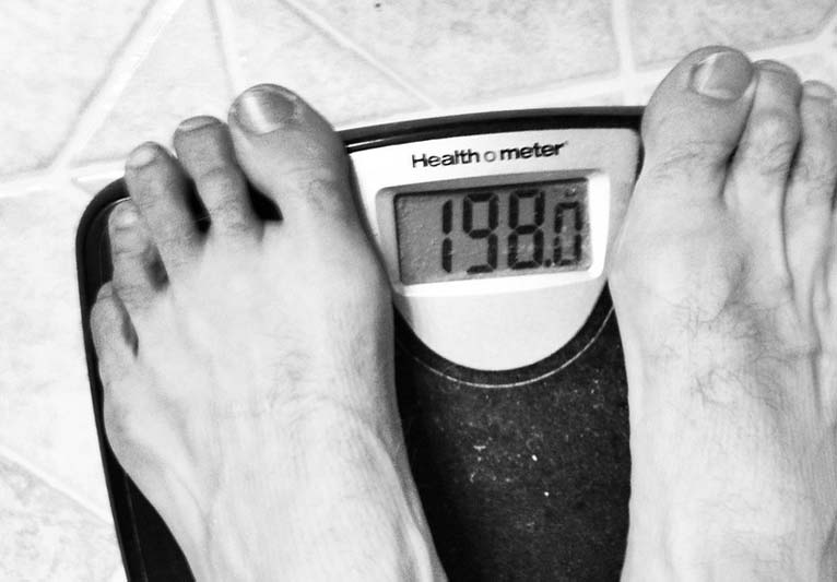 A close-up of a man's toes as he stands on a scale that reveals his weight as 198 pounds.