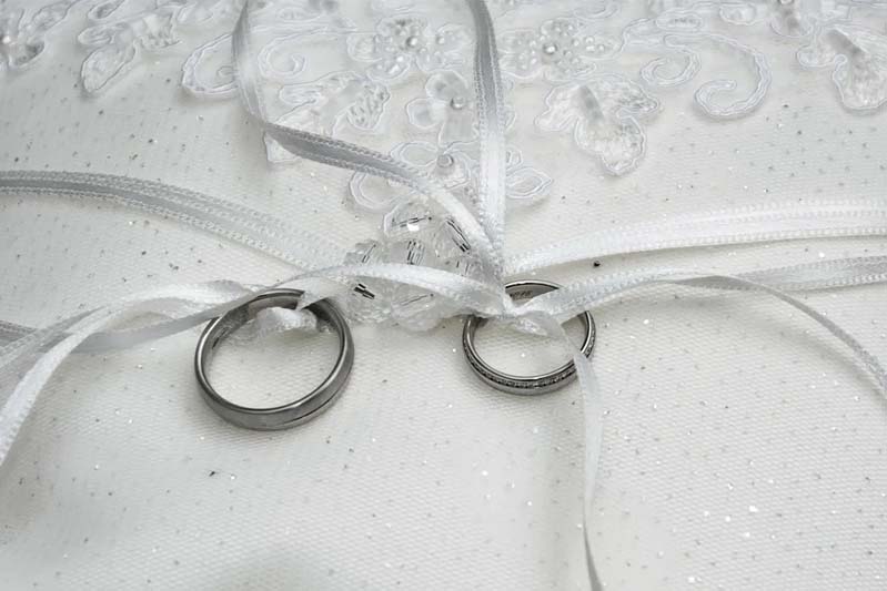 Two silver wedding rings tied to a white embroidered pillow covered in a silver sparkled tulle with white ribbon.