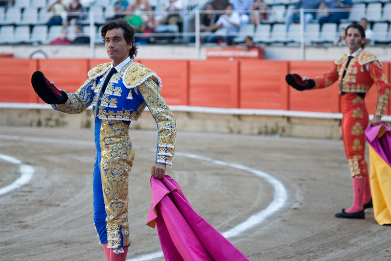 Two bullfighters in blue and gold and red and gold stand in the ring holding their hat in one hand and their pink and yellow capes in the other