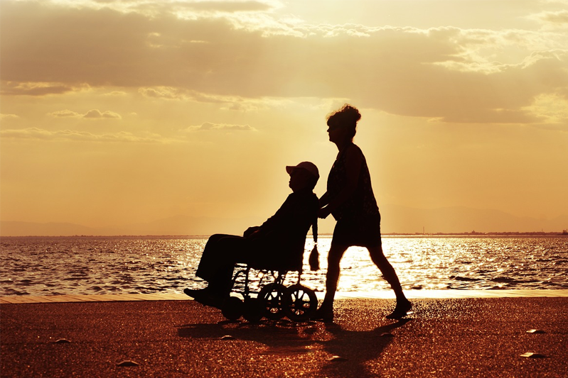 A woman pushing a man in a wheelchair along the beach at sunset.