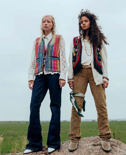 Two ladies standing on a hill in colorful vests, white shirts, jeans and brown suede pants.
