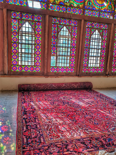 A Persian rug in primarily reds on a navy blue background has been unrolled in front of a wall of stained glass windows.