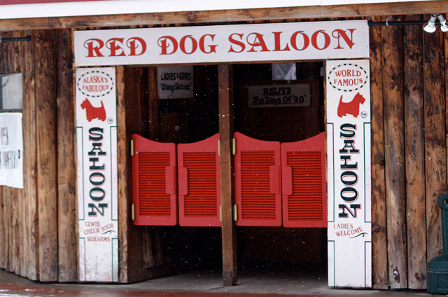 Wide white boards frame the entrance to the Red Dog Saloon with vertical wood planks on either side. The doorway has two entrances each with short red shutters.