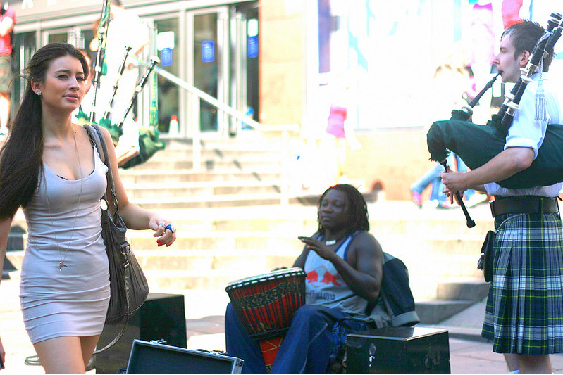 Latin woman in a tight dress, swinging her hips, as she walks on by while a black woman plays a drum and a Scotsman plays his bagpipes.