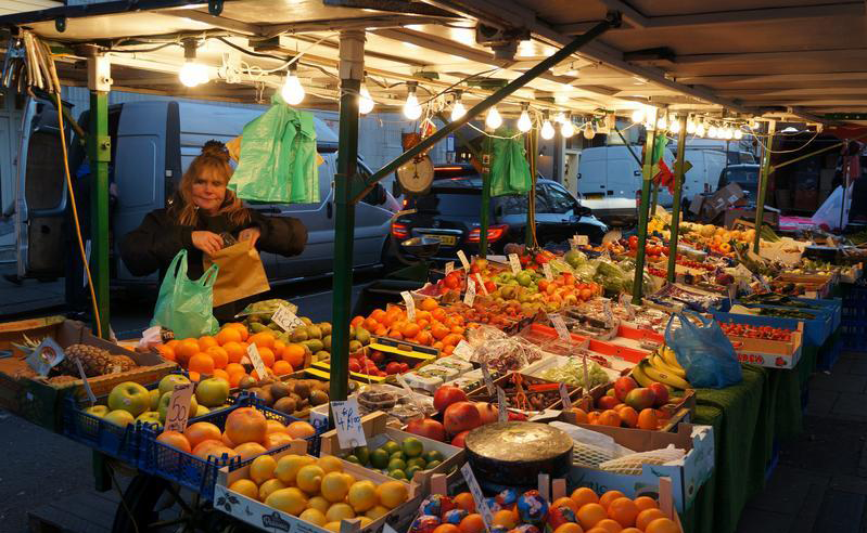 A woman stands behind long, wide tables piled with fruit
