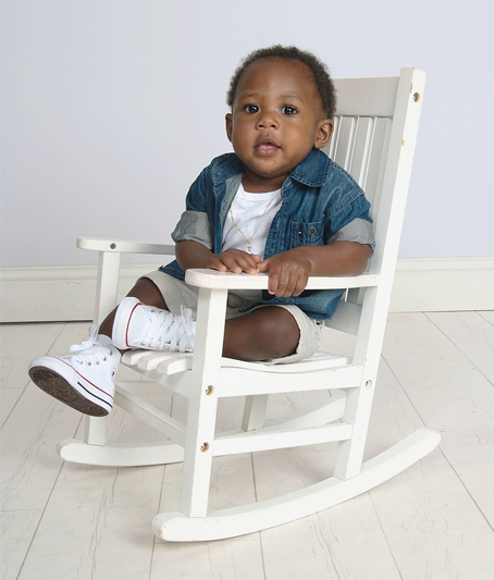 Cute little boy in white T-shirt, a denim jacket with the sleeves rolled up, pale gray shorts, and white tennis shoes is curled up in the seat of a white rocking chair set on a white plank floor, white baseboards, and a pale gray wall.