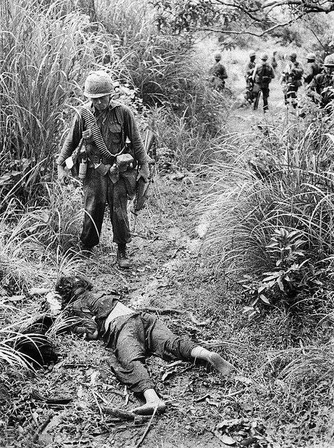 A black-and-white photo. As his men wait for him in the background, a patrol leader from a unit of the U.S. 1st Cavalry Division sorrowfully looks down at a dead Vietnamese woman.