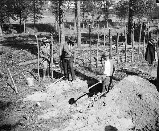 A black-and-white photo of a man with a shovel standing in a ditch while two men stand above him.