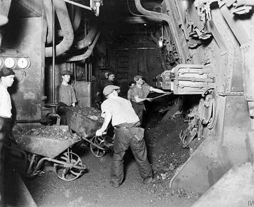 A black-and-white photo in the belly of a ship with five men either moving wheelbarrows or shoveling coal into an engine.