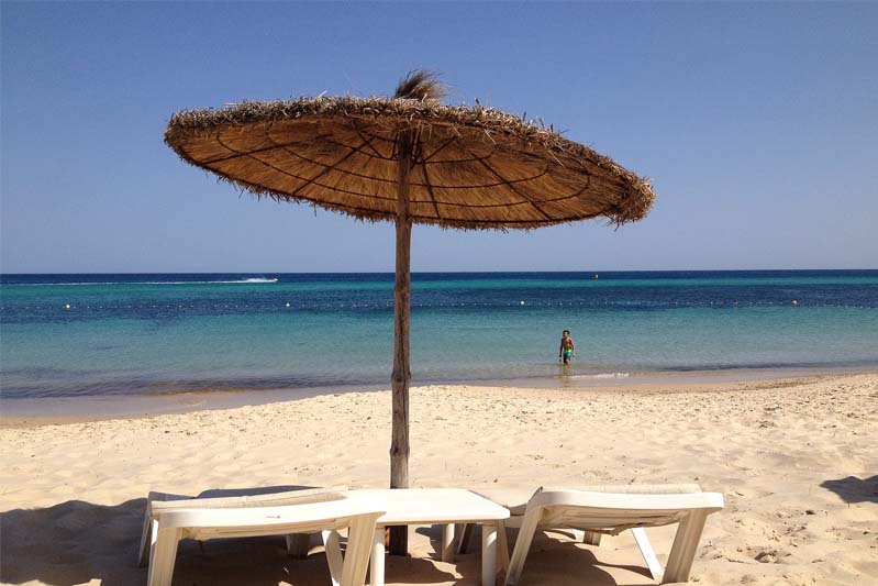 A beach with two lounge chairs under a palapa umbrella looking out over the ocean.