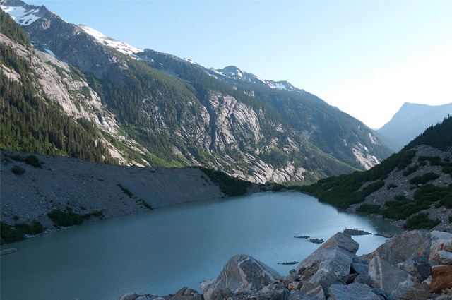 A small black body of water surrounded by tall mountains with boulders in the right foreground.