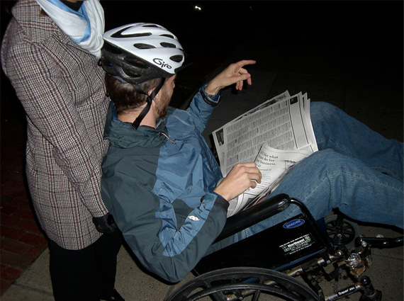 Woman in a brown plaid coat wearing a white and blue scarf is pushing a man wearing a two-tone blue windbreaker, jeans, and a white bike helmet in a wheelchair, who is pointing somewhere..