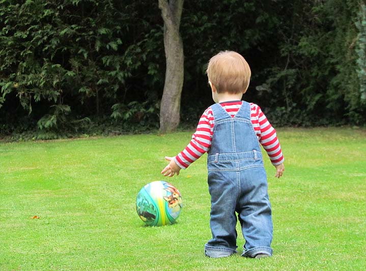 Toddler in overalls and red-and-white striped shirt walking towards a colorful ball on green grass.