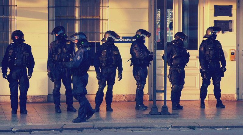 A line of riot ready cops in front of a yellow building.