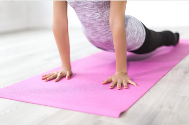 A three-quarter profile of a woman in a streaky grey top and black yoga pants pushing up with her arms on a pink yoga mat.