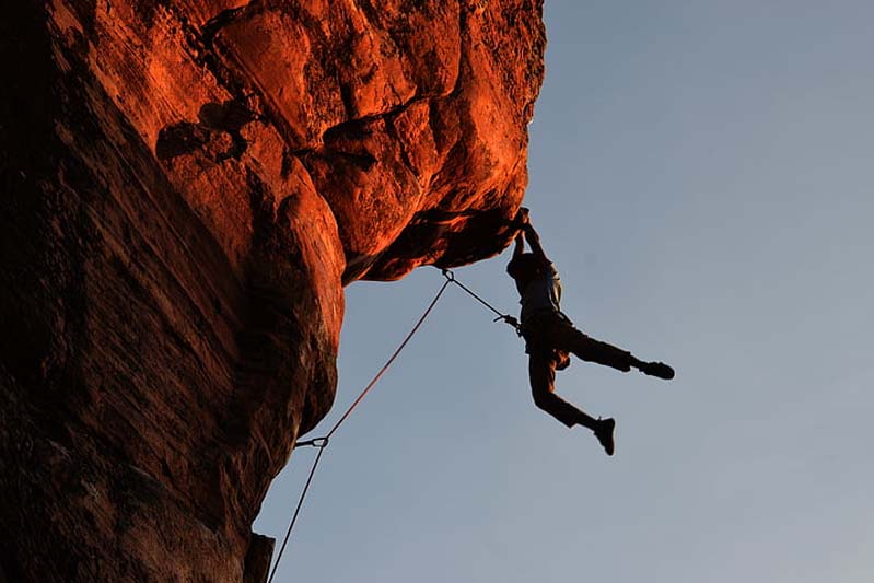 A climber hanging under a jutting red-orange rock against a gray sky.