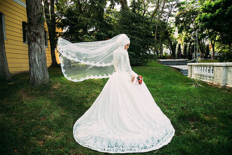 A bride in white dress and veil is twirling around on a lawn with a yellow house.