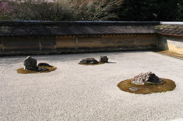 A zen garden of raked sand, the clay wall is stained by age with subtle brown and orange tones and three isolated rounds of grass with rocks in their centers.