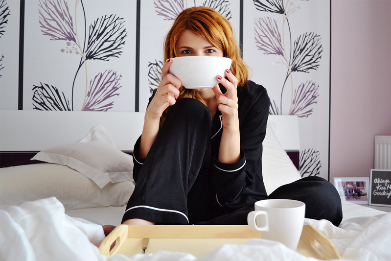 Woman in black pjs sitting up in bed with a white bowl in her hands and a cup of coffee on a tray