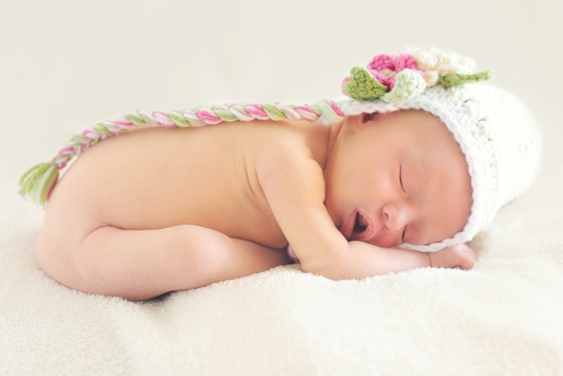 Naked baby all curled up on her stomach on a sherpa blanket, fast asleep while wearing a white knit cap with a big pink flower and a pink, white, and green twisted braid for a tie