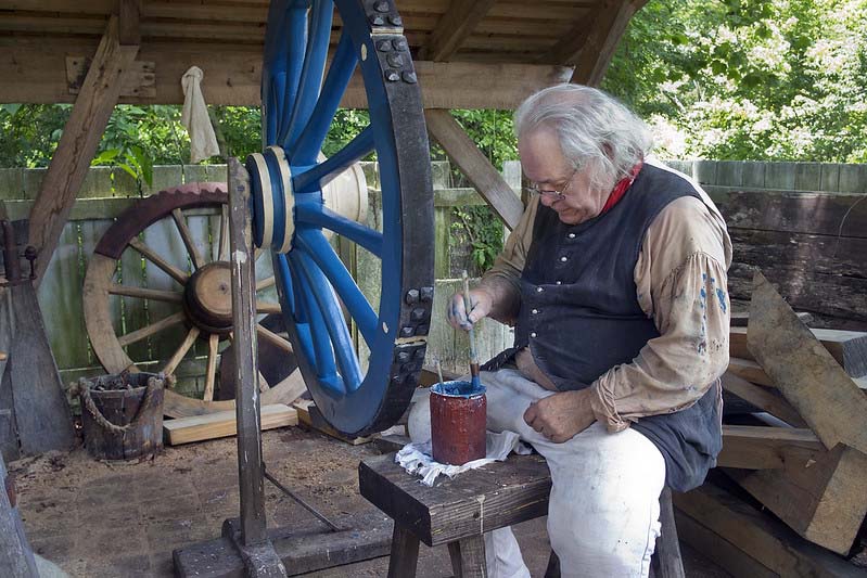 A costume gentleman is sitting on a wooden bench dipping a brush into a paint pot of blue, painting a wooden wheel in a reproduction shop.