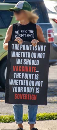 A blonde woman in a white and green baseball cap and black tanktop and jeans holds a black rectangular sign outside a parking lot.