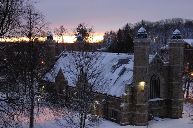 An aerial view at twilight of a small chapel covered in snow and surrounded by snow-draped trees.