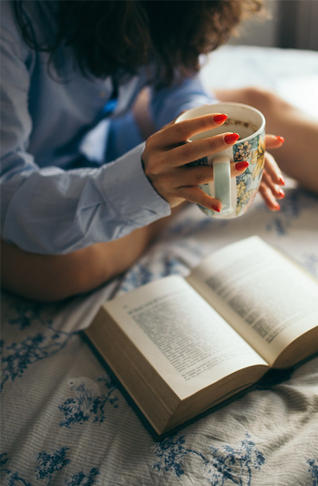 Dark-haired woman in a blue button-down shirt holds a cup of coffee in her hands as she reads a book sitting on a bed with a tan and white striped bedspread with blue flowers 
