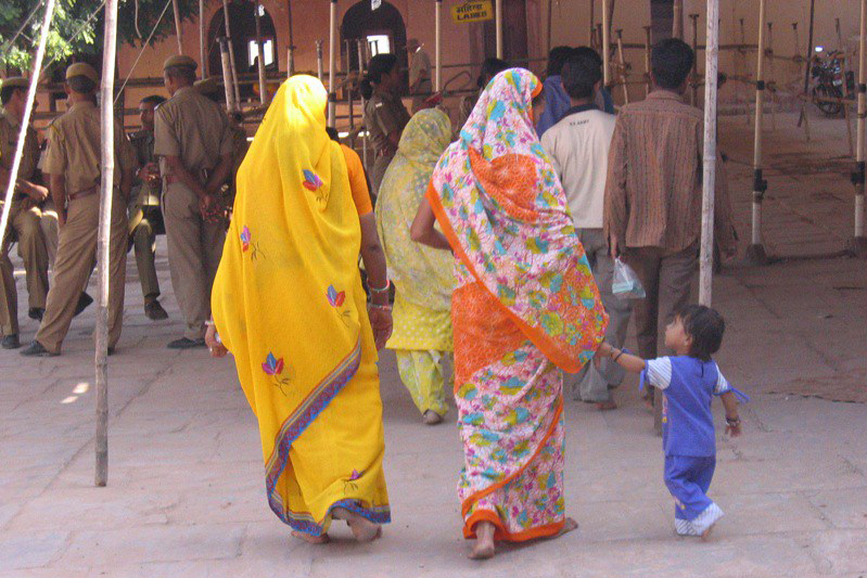 Two women walking away from us are wrapped in a yellow sari (on the left) and an orange sari (on the right) with a wide lavendar and blue floral border, as she holds her child's hand.